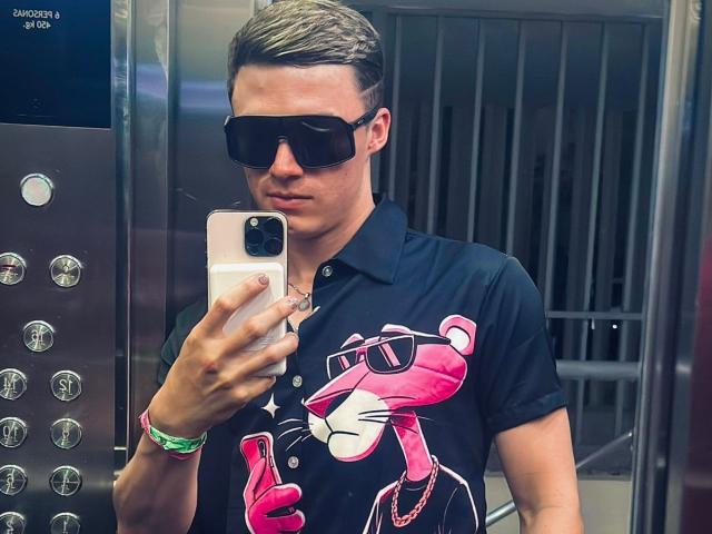 ErosHot with short brown hair, black sunglasses, and a pink cartoon panther shirt, holding a phone, looking at it, wearing a green bracelet, in a mirror selfie.