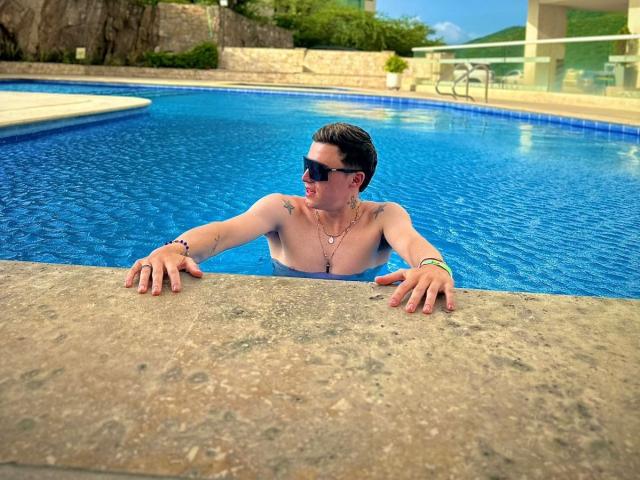 ErosHot, shirtless with tattoos, wearing dark sunglasses, leans on a pool edge, water up to chest, hands on concrete, blue pool in background, sunny day, relaxed.