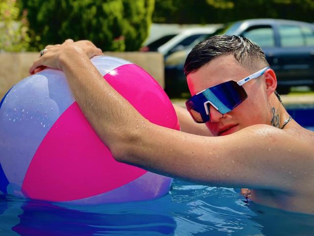 ErosHot with short dark hair, blue mirrored sunglasses, and a tattoo, holding a pink and purple inflatable ball in a blue swimming pool. Water droplets on his skin.