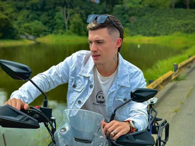 ErosHot with short brown hair, white denim jacket, and sunglasses on head, riding a motorcycle, focused, wearing a white tee, right hand on handle, left hand on brake.