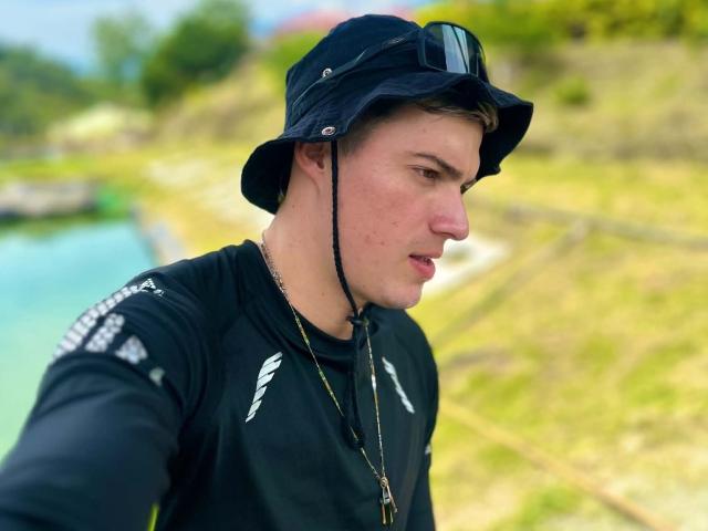 ErosHot, a young white guy with short brown hair, wearing a black bucket hat and wetsuit, looking to the side with a slight frown. His wetsuit has white patterns.