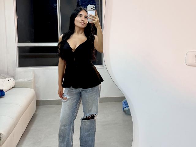 EmilyJensenn with long black hair, black cleavage-cut top, and ripped blue jeans, taking a mirror selfie, showing her tits and ass, standing in a white room.
