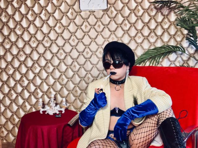 FemCupid with black hair, sunglasses, blue gloves, yellow check coat, black bra, fishnet stockings, black boots, sucking on black lollipop, sitting on red couch, arms crossed.