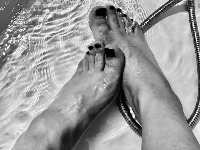 FemCupid's wet feet with black-painted toenails, crossed at the ankles, showing water droplets and a metal shower hose in a black-and-white photo.