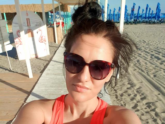 JoliDanielle with dark hair in a messy bun, wearing red sunglasses and a red top, showing her pouty face and sunlit skin, standing on a sandy beach.