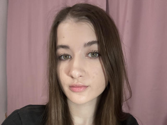MisscharyNessy has fair skin, green eyes, and brown hair, with a slight pout and shiny pink lips, wearing a black shirt, against a pink curtain.