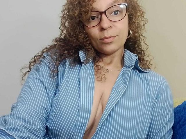 Angelonix with curly brown hair, glasses, and a blue striped shirt, slightly open to show her large, natural, bare tits. She has a neutral expression and a silver necklace.