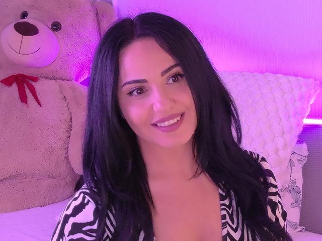 Jenniferwild69 with long black hair, smiling, wearing a zebra-patterned top, purple lighting, large beige teddy bear with red ribbon, white textured pillow, and gray patterned pillow.
