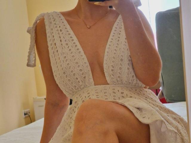 Jumiera in a white, deep V-neck, lace dress, showing her tits and a thigh in a mirror selfie, holding a phone, with a gold necklace.