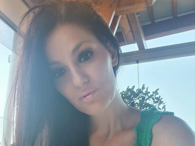 JoliDanielle with long brown hair, green eyes, and shiny pink lips, wearing a green top, has a cute, sexy face, and a small beauty mark on her right cheek.