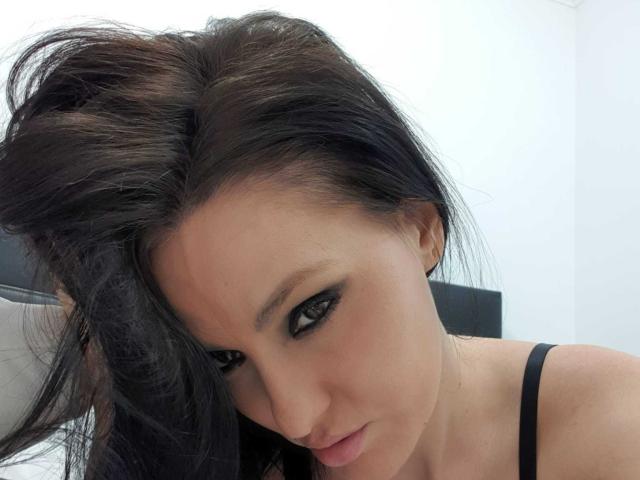 JoliDanielle with dark, wavy hair, black eyeliner, and a slight smirk, wearing a black strap, looking directly at the camera with a sexy, confident expression.