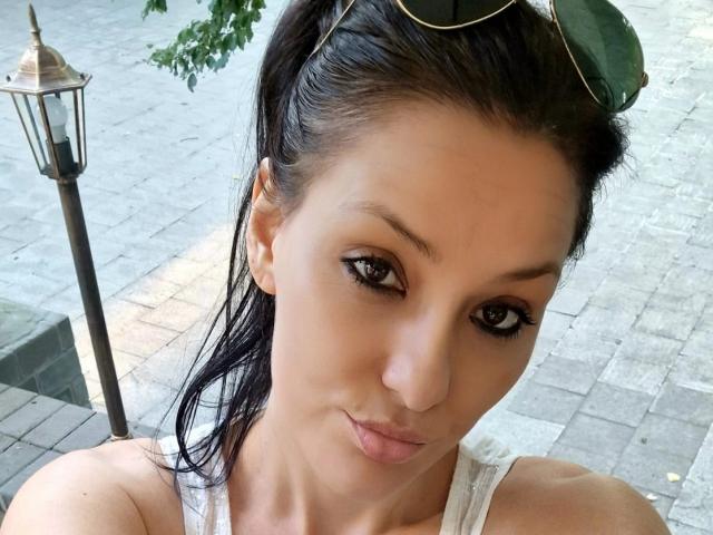 JoliDanielle with pouty lips, black hair in ponytail, wearing sunglasses on head, green eyes, and white tank top, showing off her small tits. Lamp in background.