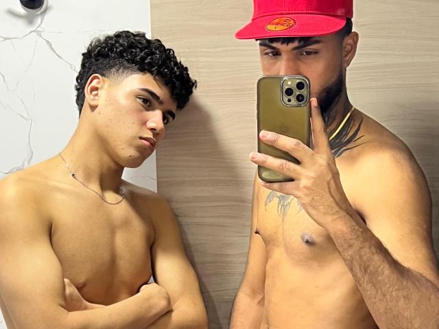 Two shirtless ImperialMen, one with curly black hair and tan skin, arms crossed, looking side. The other, with a red cap, tattoos, and brown skin, taking a mirror selfie.