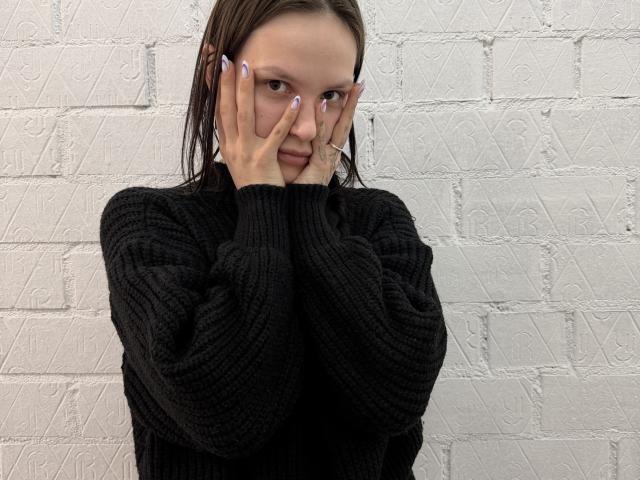 AlaneHarradon has wet brown hair, wearing a black ribbed sweater, hands covering her face with purple nails, cigarette in right hand, looking serious, standing against a white brick wall.