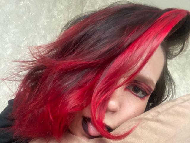 AlaneHarradon has red and black hair, black lipstick, red eyeshadow, and a pale face, with a pillow under their chin, looking at the camera with a slightly open mouth.
