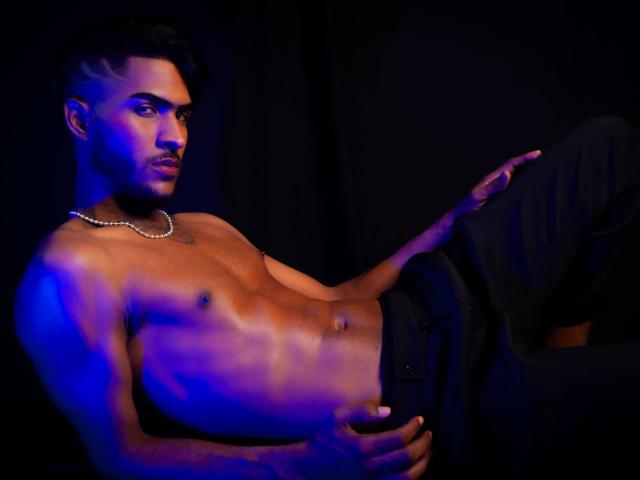 TheImperialMen, shirtless with blue lighting, wearing black pants and a pearl necklace, laying back, hand on pants, muscular, dark hair, slight beard, serious expression, black background.