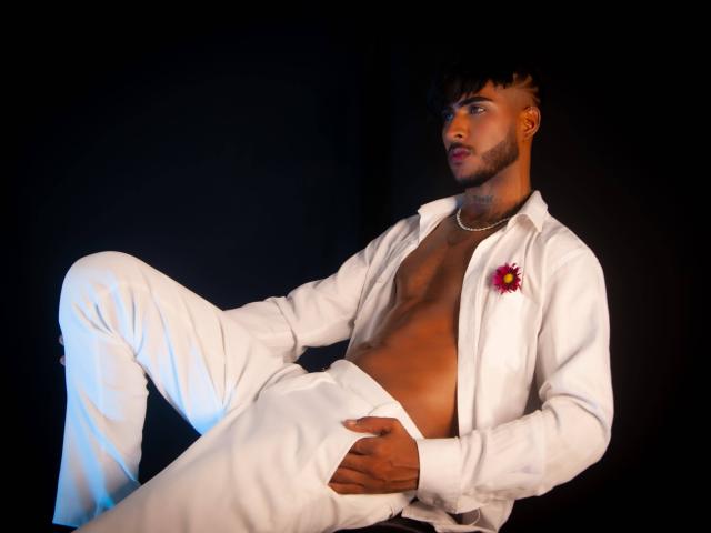 TheImperialMen, a dark-skinned dude with a beard, wearing a white shirt and pants, open to show his chest, with a flower on the shirt, leaning back.