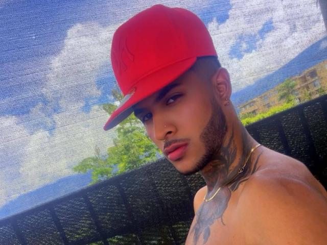 TheImperialMen has a red cap, tattooed neck, and chest, with a serious look, shirtless, outdoors, with a blue sky and white clouds reflected in the mesh.