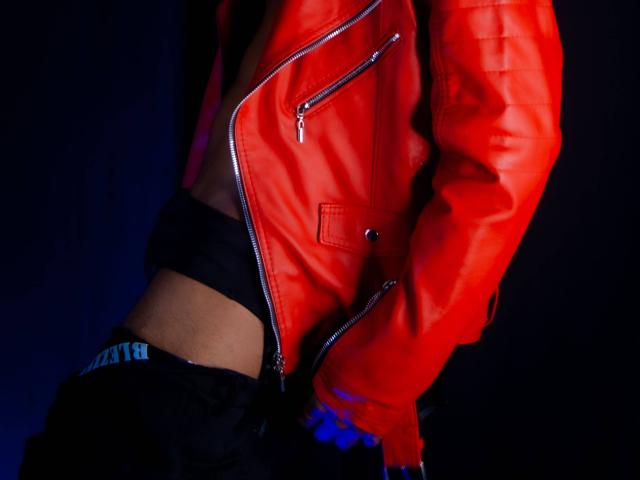 TheImperialMen wearing a bright red leather jacket with zippers, black pants, and blue gloves, showing a slim, toned midsection. Hands in pockets, dark background.