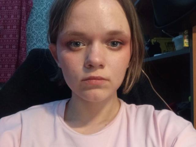 AngeliaAmstutz has a tired, puffy face with blue eyes, wearing a pink shirt, looking slightly to the side with a serious expression, and brown hair.
