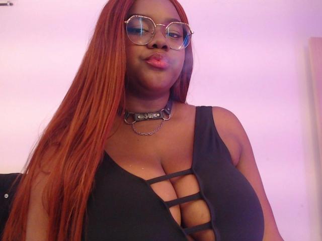 RoseBeckers with long red hair, wearing glasses, black strappy top showing large tits, and a metal choker. She's pouting with pink lipstick.