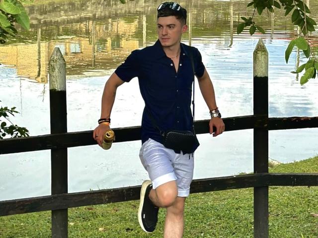 ErosHot with short dark hair, wearing a navy shirt, white shorts, black sneakers, and a black crossbody bag, leans on a wooden fence with a beer in hand.