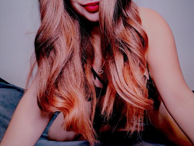 GiannaSexxy with long, wavy, pink-to-brown ombre hair, red lipstick, black strap dress, heart necklace, and visible tits, sitting on a grey blanket.