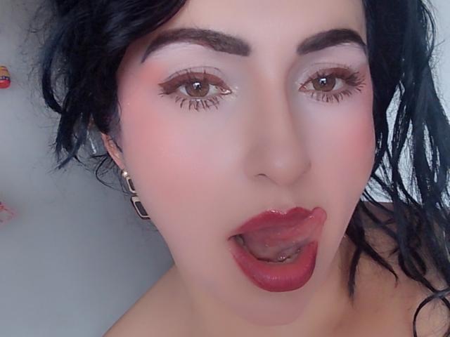 LuluFrancaise has dark wavy hair, brown eyes, and is sticking out her tongue with red lipstick and a small lip piercing. She's wearing bold eyeliner and a gold earring.