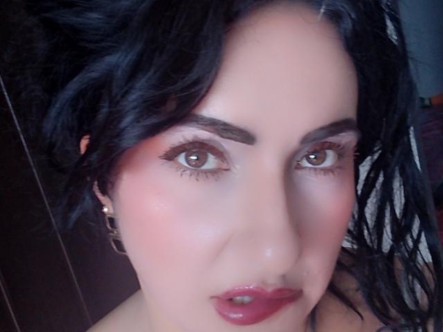 LuluFrancaise has dark wavy hair, brown eyes, and red lipstick. Her face is slightly flushed with pink makeup. She wears gold earrings. Her lips are slightly parted.
