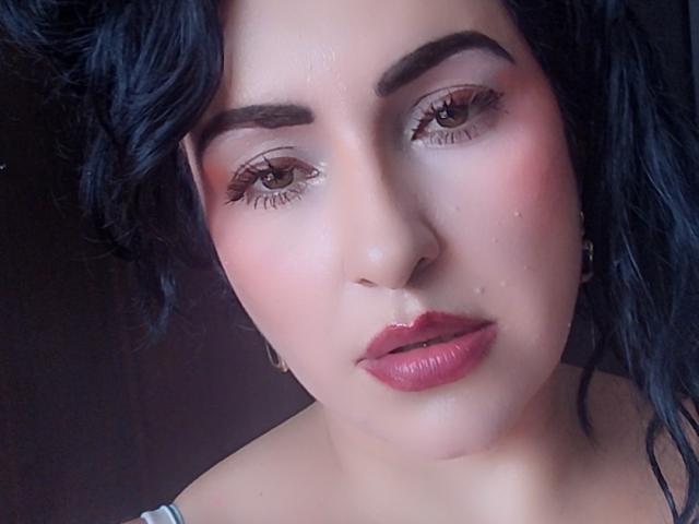 LuluFrancaise has dark curly hair, brown eyes, and pink glossy lips with a slight sheen on her face. Her eyebrows are thick and she's wearing subtle makeup.