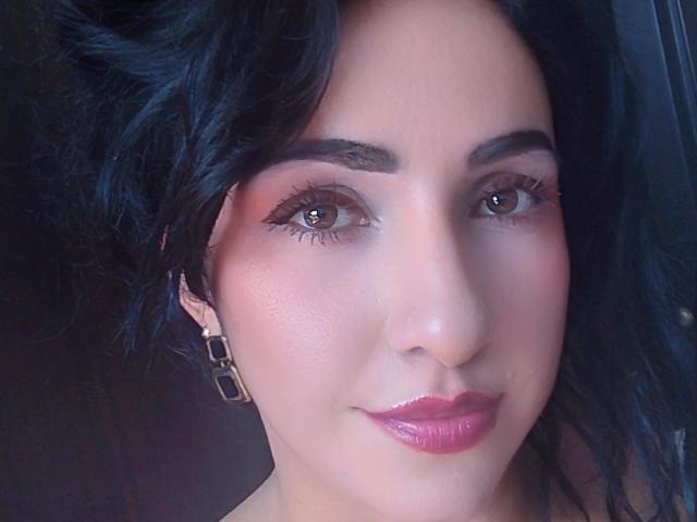 LuluFrancaise's face, with dark wavy hair, brown eyes, pink lipstick, and shiny pink lips, wearing square earrings, looking straight at the camera.