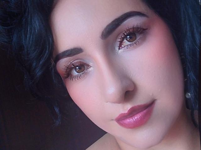 LuluFrancaise has a pretty face with pinkish blush, dark wavy hair, thick eyebrows, brown eyes, and pink lipstick. Her boobless chest is partially visible in the top frame.