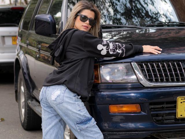 SheylaSpooner leans against a dark blue SUV, wearing a black hoodie with white designs, and ripped blue jeans, with her ass slightly raised and sunglasses on.