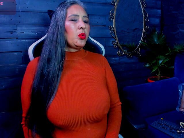 VictoriaKiley with long black hair, red lipstick, and orange ribbed sweater, pouting her full, rounded, and large, firm tits, sitting in a blue wooden room.