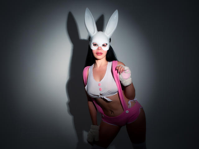 HelenHard with white bunny mask, pink shorts, white crop top, pink suspenders, white gloves, and pink wristbands, showing her tits and ass, shadowed against a dark background.