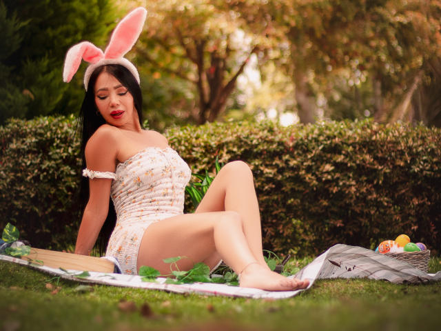 HelenHard with pink bunny ears, white off-shoulder dress, and red lipstick, sitting on a blanket, showing her ass and bare feet, with Easter eggs and a basket nearby.