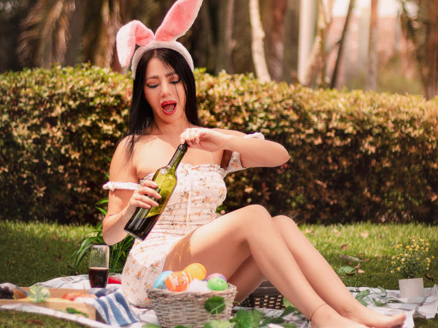 HelenHard with pink bunny ears, pouring wine from a bottle, sitting on a blanket with Easter eggs, striped blanket, and a glass of dark liquid. She's wearing a floral dress.