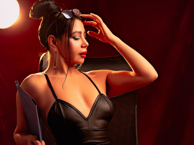 HelenHard with brown hair in a bun, black leather dress, holding clipboard, adjusting glasses, red lipstick, sitting in chair, sexy, holding clipboard in left hand, right hand on head.