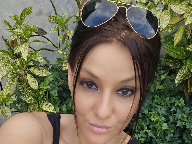 JoliDanielle has dark brown eyes, light brown skin, and wears black eyeliner. She has her brown hair up, sunglasses on her head, and a pouty expression. Her tits are small.