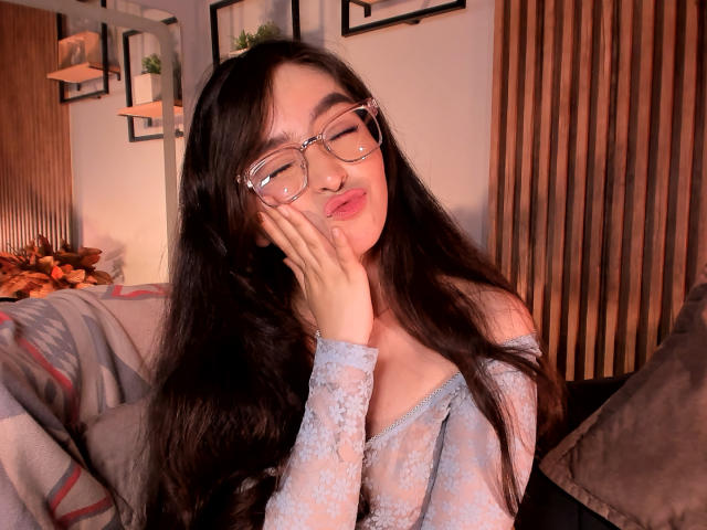 EvaMcAllister with long black hair, glasses, and pouty lips, touching her cheek, wearing a sheer blue lace off-shoulder top, showing her small tits. Bedroom setting, soft lighting.