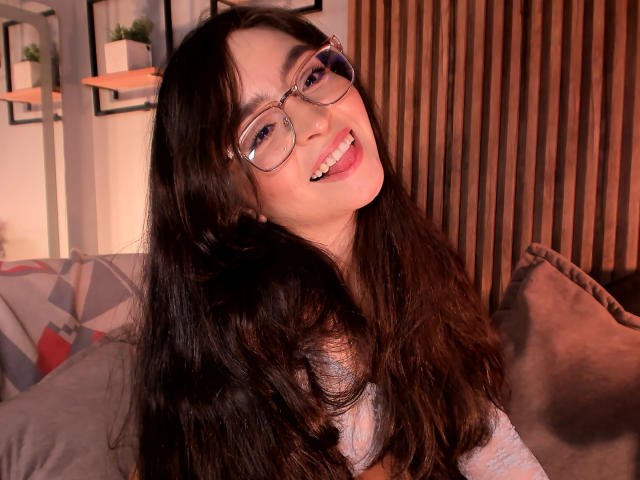 EvaMcAllister with long, wavy dark hair, glasses, and a cute smile, sticking her tongue out. She's wearing a light gray top, looking happy and playful.