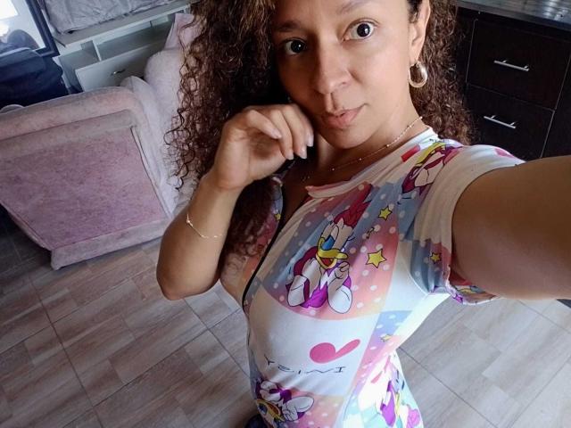 Angelonix with curly brown hair, wearing a colorful Winnie the Pooh dress, gold hoop earrings, and a gold bracelet, taking a selfie, showing her chest.