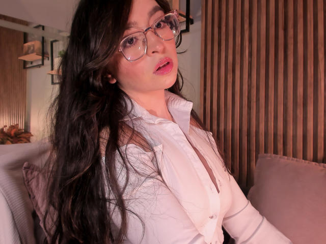 EvaMcAllister with long black hair, glasses, pink lipstick, and white button-up shirt, looking at camera, showing her tits, in cozy indoor setting, wooden wall in background.