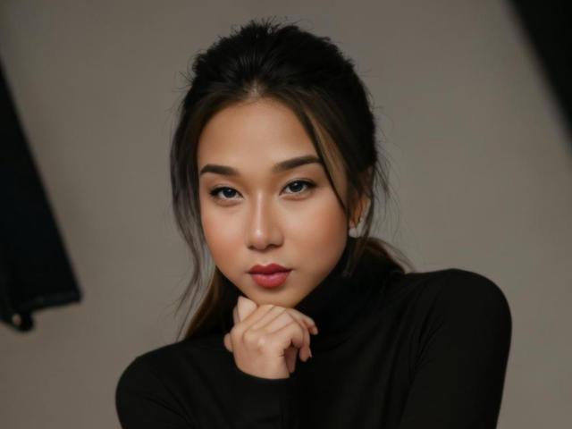 OneSeductiveDollTS with dark hair, wearing a black turtleneck, resting chin on hand, looking at camera with subtle smile, soft makeup, and dark eyebrows, in a relaxed pose.