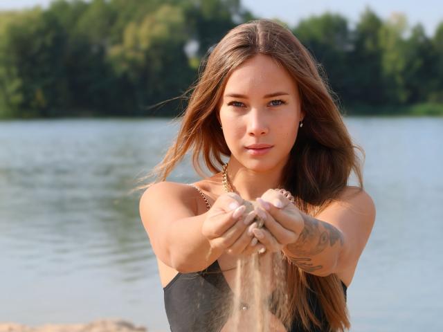 AmmaLoyXX with long brown hair, blue eyes, and tattoos on her right arm, pouring water from her hands, wearing a black tank top, standing by a lake.