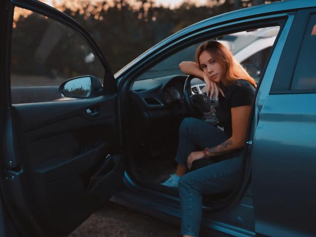 AmmaLoyXX with orange hair sits in a blue car, leaning on the door with her arm, wearing a black top and jeans, looking at the camera. Tattoo on her arm.
