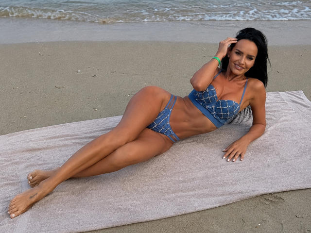 SexyGirl with tan skin, black hair, and small tits, lying on a towel in blue plaid bikini, smiling, hand in hair, beach, legs stretched out, water in background.