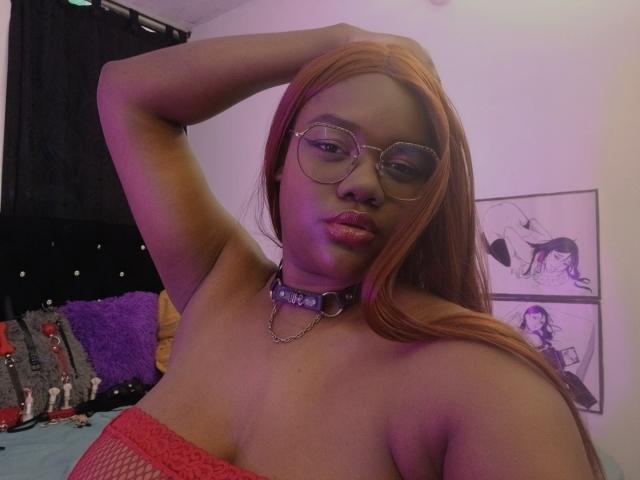 RoseBeckers with long orange hair, glasses, and a purple choker, wearing a red fishnet top, raising her arm, showing her tits, and sucking on her lip.