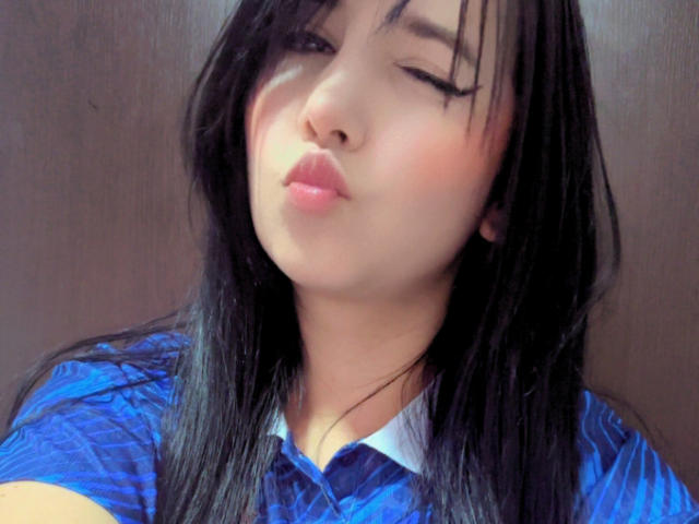 Kkatalina with black hair, winking, and pouting her plump, shiny pink lips, wearing a blue shirt with white collar, against a dark background.
