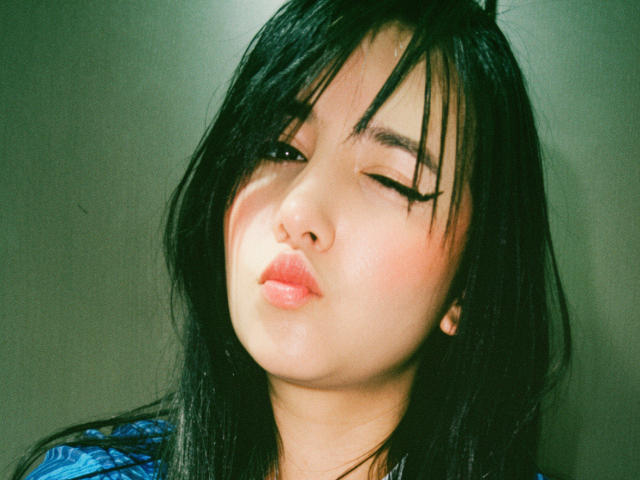 Kkatalina's puffy pink lips are slightly parted, eyes half-closed with long black eyelashes, black straight hair covering her forehead, and a soft greenish light on her fair skin.